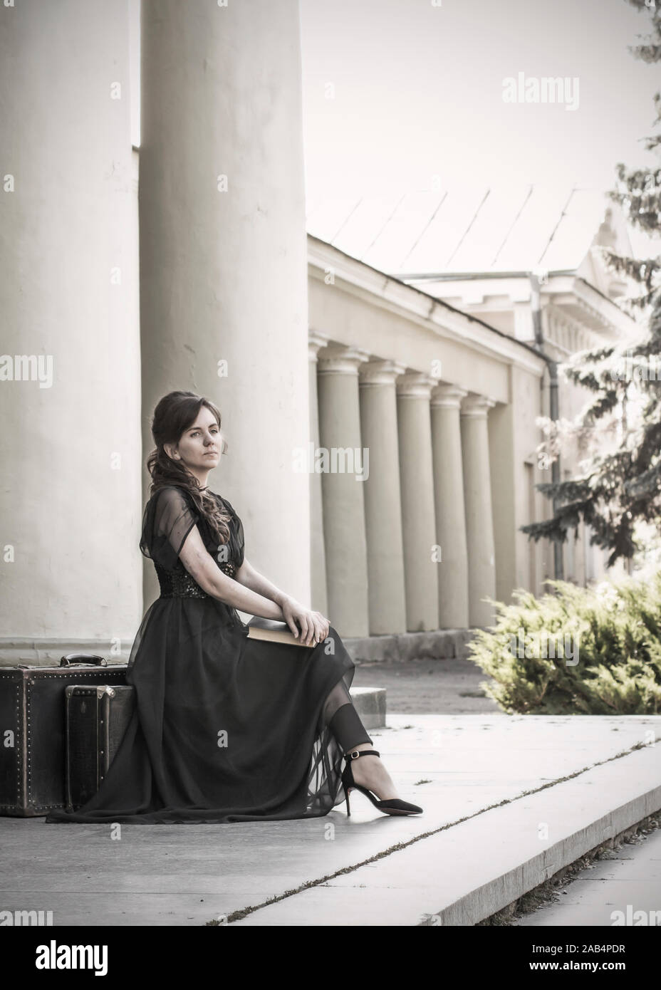 Schöne Mädchen in einem schwarzen Abendkleid sitzt mit einem Buch. Ein Mädchen sitzt mit Koffern in der Nähe ein altes Gebäude mit Säulen. Schöne Frau mit Narben. Re Stockfoto