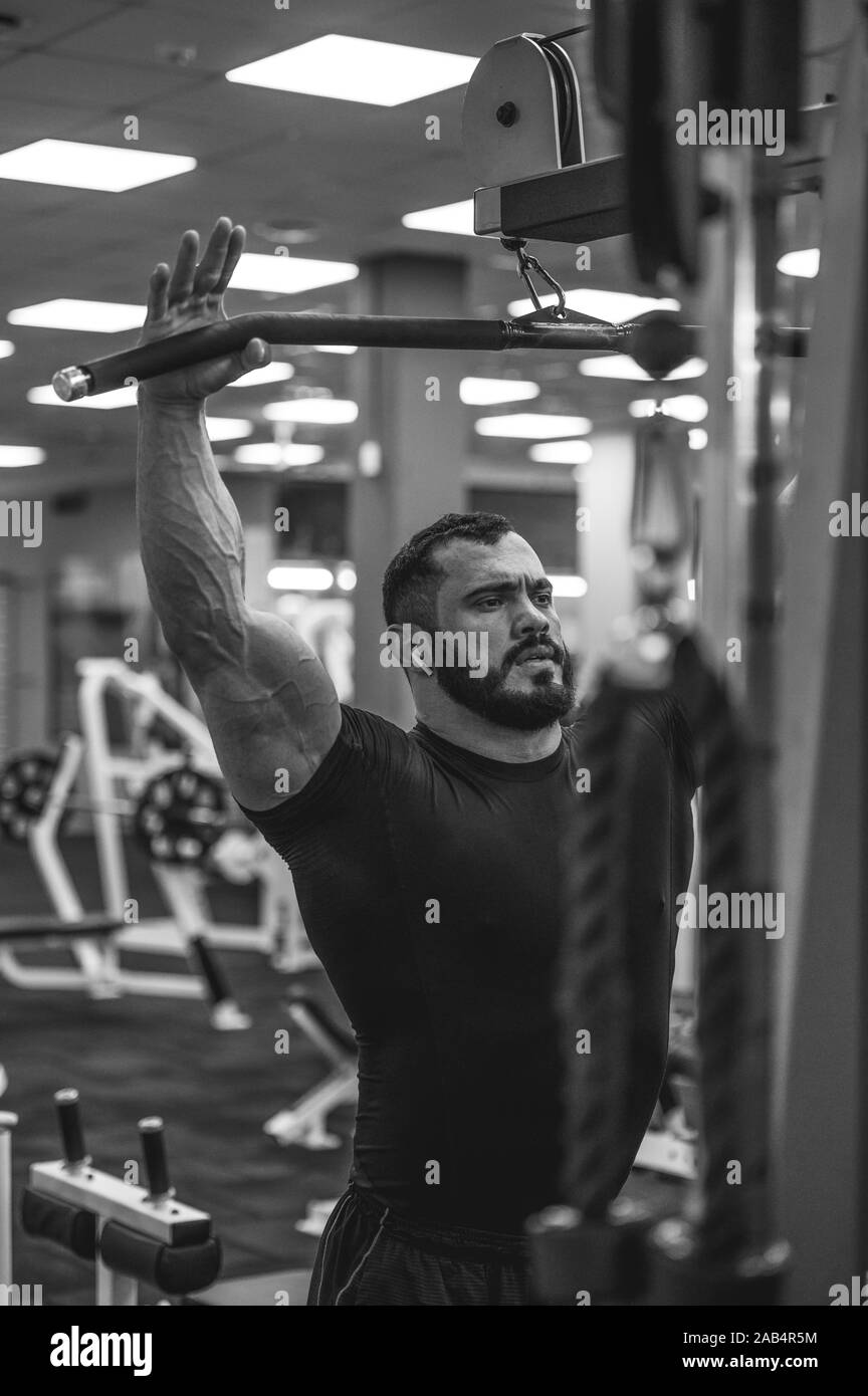 Junge starke kaukasischen bärtiger Mann im schwarzen T-Shirt mit drahtlosen Kopfhörern in den Ohren Ziehen von schweren Gewicht auf Fitnessgeräte indoor Sport Fitnessraum schwarz ein Stockfoto