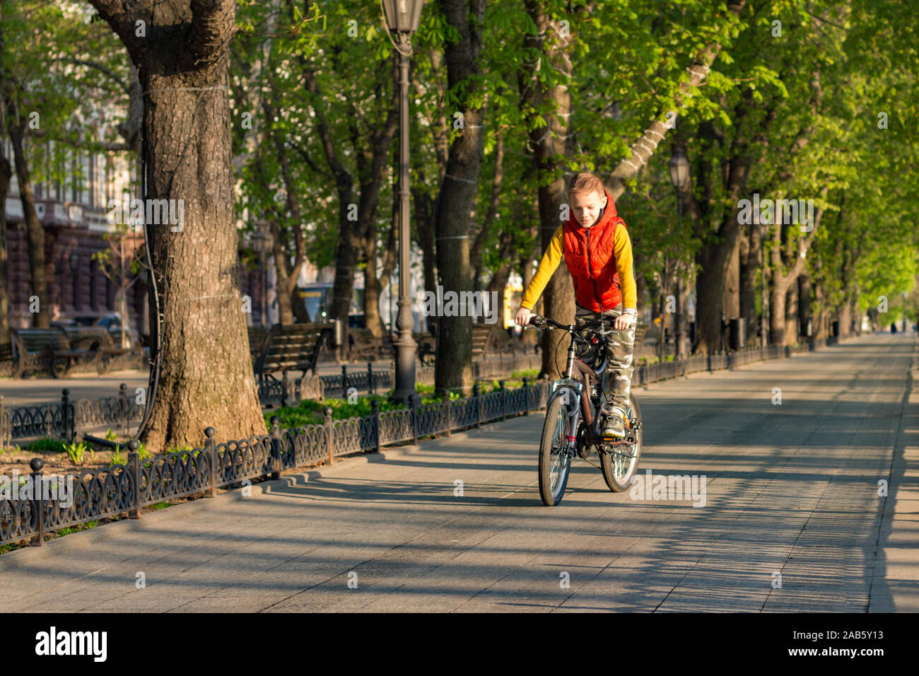 Ein Junge von 8 Jahren in einem orange Weste reitet ein Fahrrad in einem Stadtpark an einem sonnigen Frühlingsmorgen Stockfoto