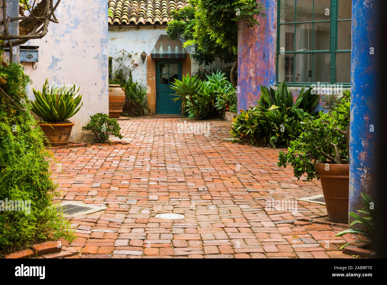 Malerische Garten im Innenhof mit Topfpflanzen und durch rustikal verputzten Wänden in lebendigen Farben, lila, pink und blau und mit Ziegeln gepflasterten Stockfoto