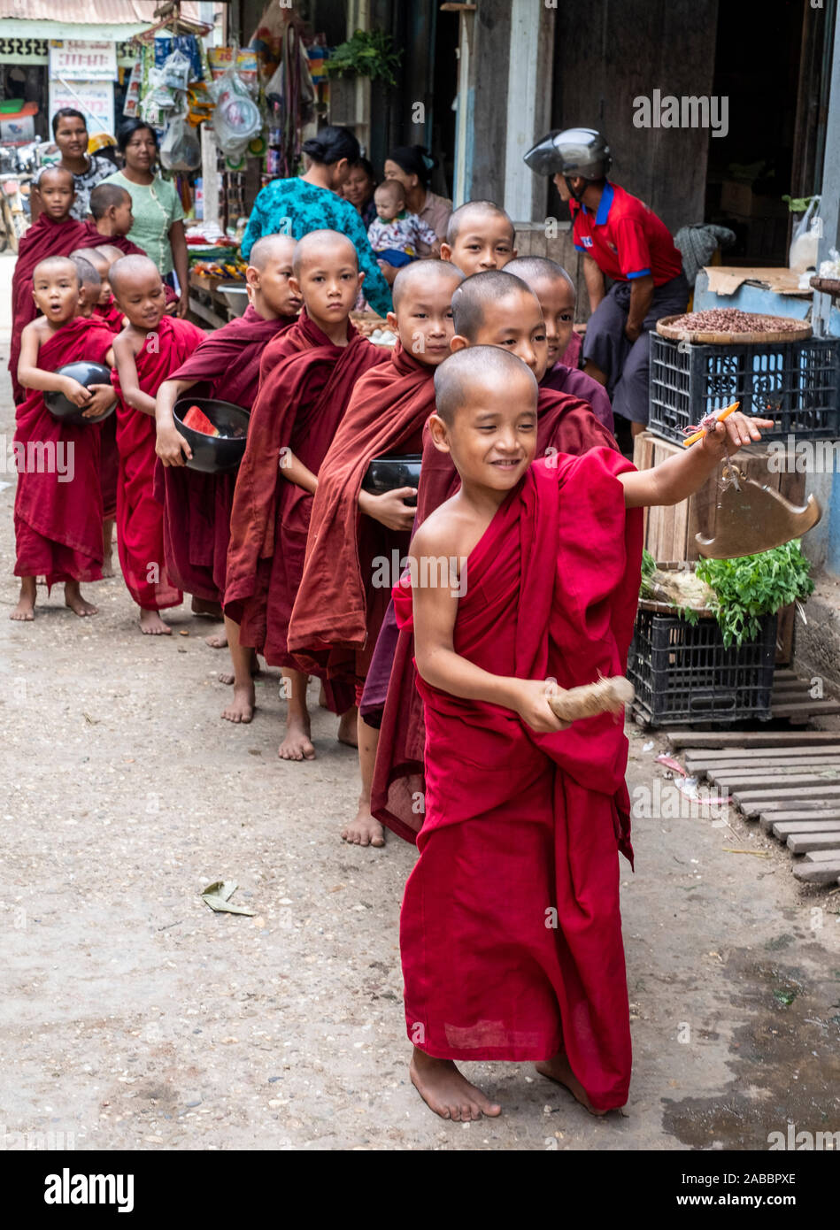 Viele junge buddhistische Mönche in roten Roben gekleidet geben Sie den Markt der Kanne, Myanmar (Birma) auf der Suche nach Almosen und von einem jungen Mönch, der mit einem Gong led Stockfoto