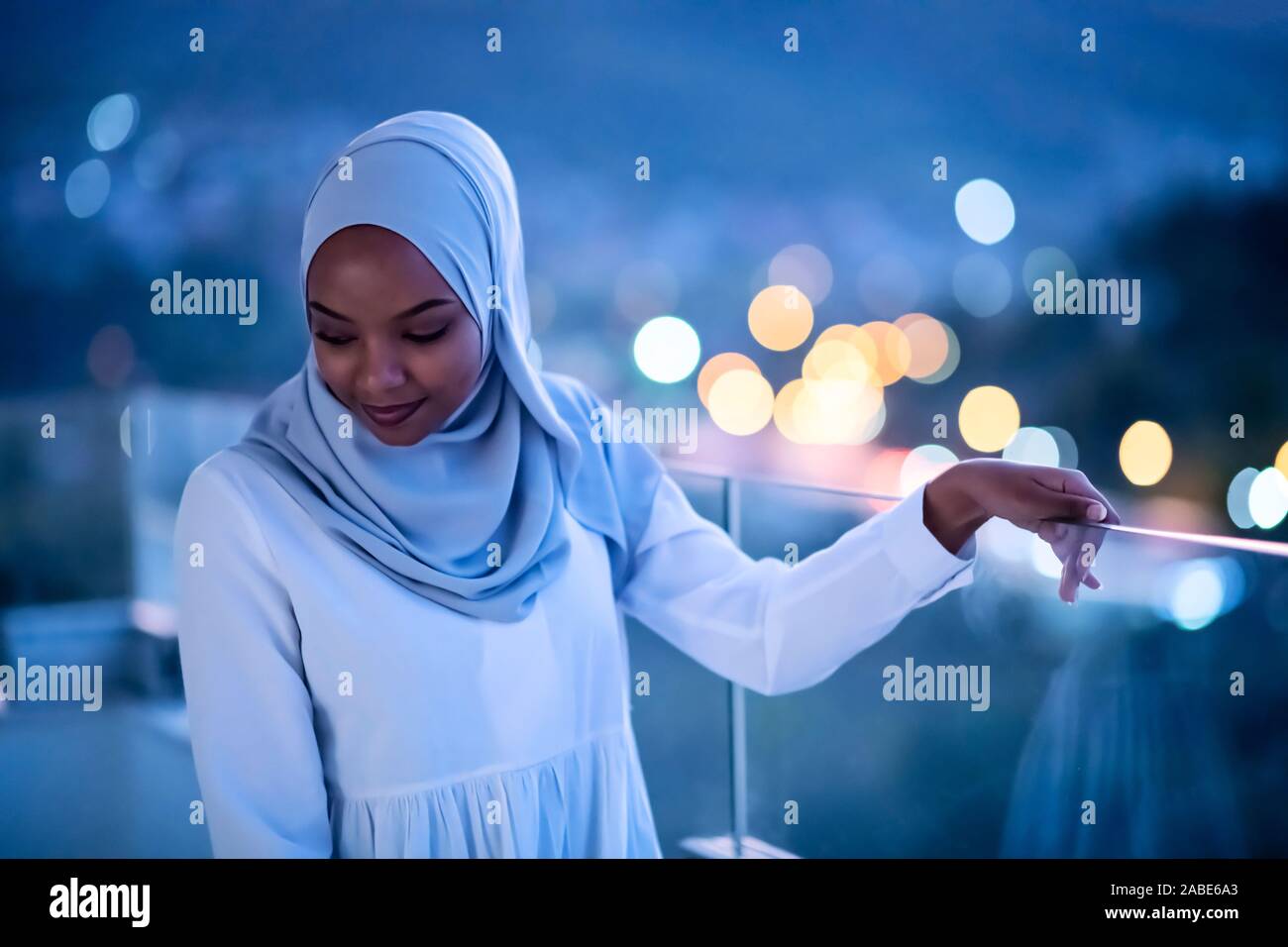 Afrikanische Moderne muslimische Frau in der Nacht am Balkon lächelnd an Kamera mit Stadt bokeh Lichter im Hintergrund Stockfoto