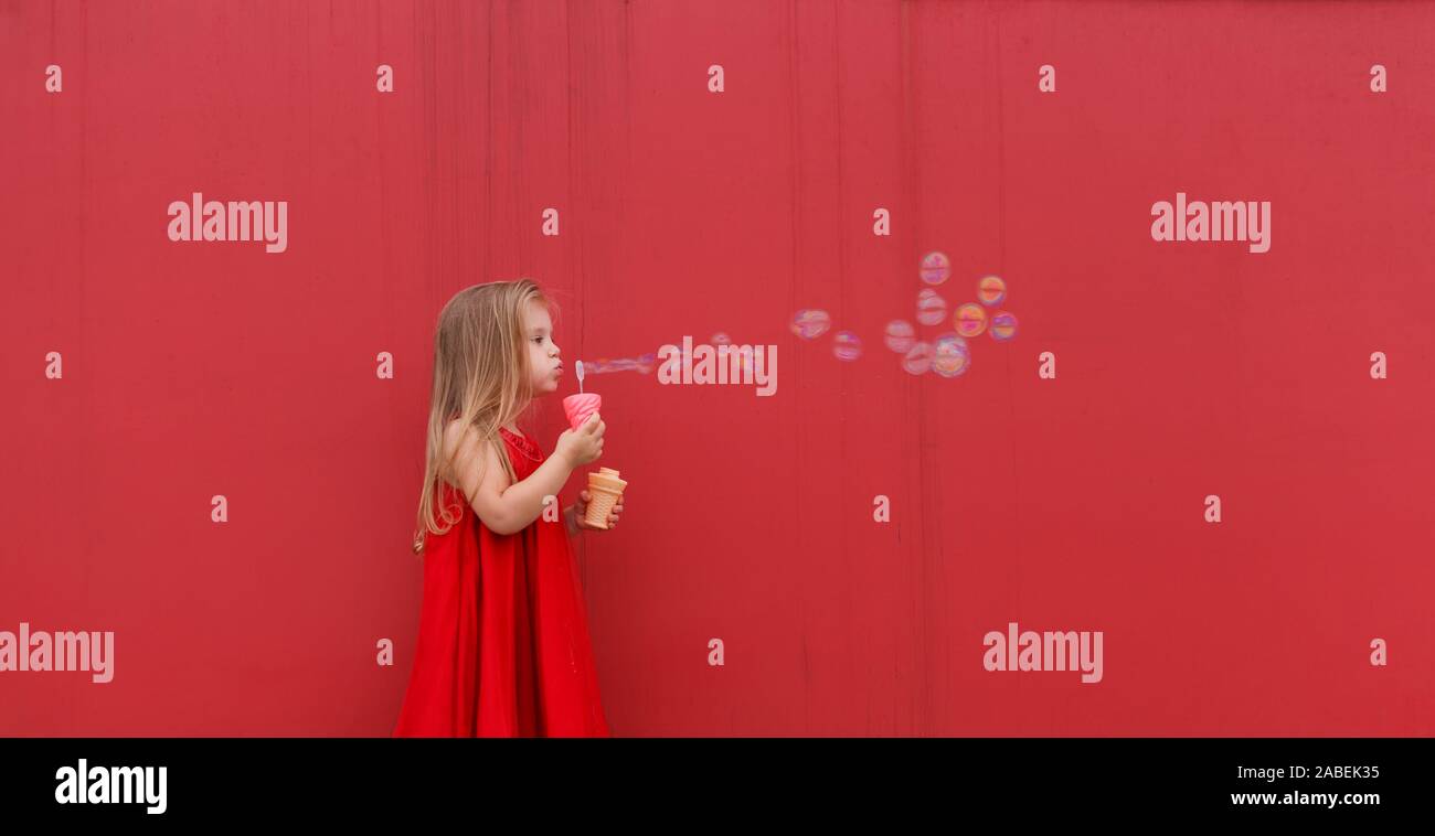 Blonde Mädchen im roten Kleid auf einem Hintergrund von der Wand bläst Seifenblasen. Stockfoto