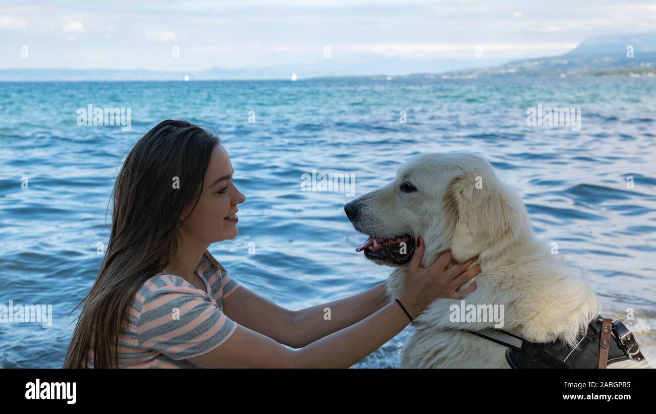 Lächelnde junge Frau mit hellbraunem Haar hält einen großen, weißen Hund auf einen unscharfen Hintergrund der Blaue See und die Berge. Stockfoto