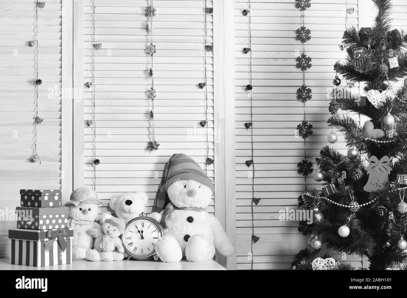 Schneemänner und Teddybären in der Nähe der Uhr und Weihnachtsbaum. Spielzeug auf Kommode auf Holz Wand Hintergrund. Tannenbaum mit Boxen und Dekorationen. Feiern und das Neue Jahr Einrichtung Konzept Stockfoto