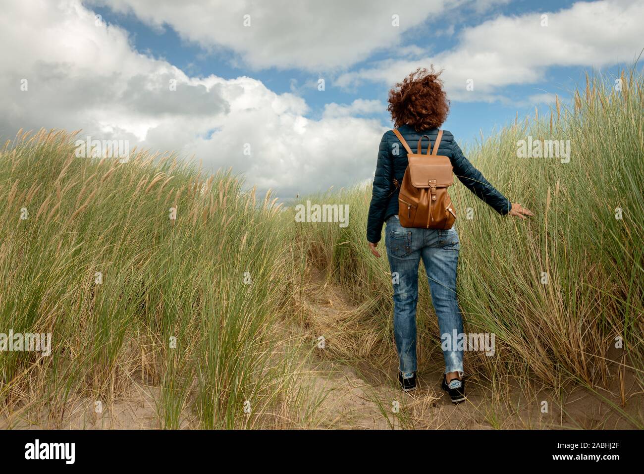 Model entließ eine junge Frau mit roten Locken, die eine legere blaue Jacke und Jeans trägt und auf einer Sanddüne durch Marram Gras wandert Stockfoto