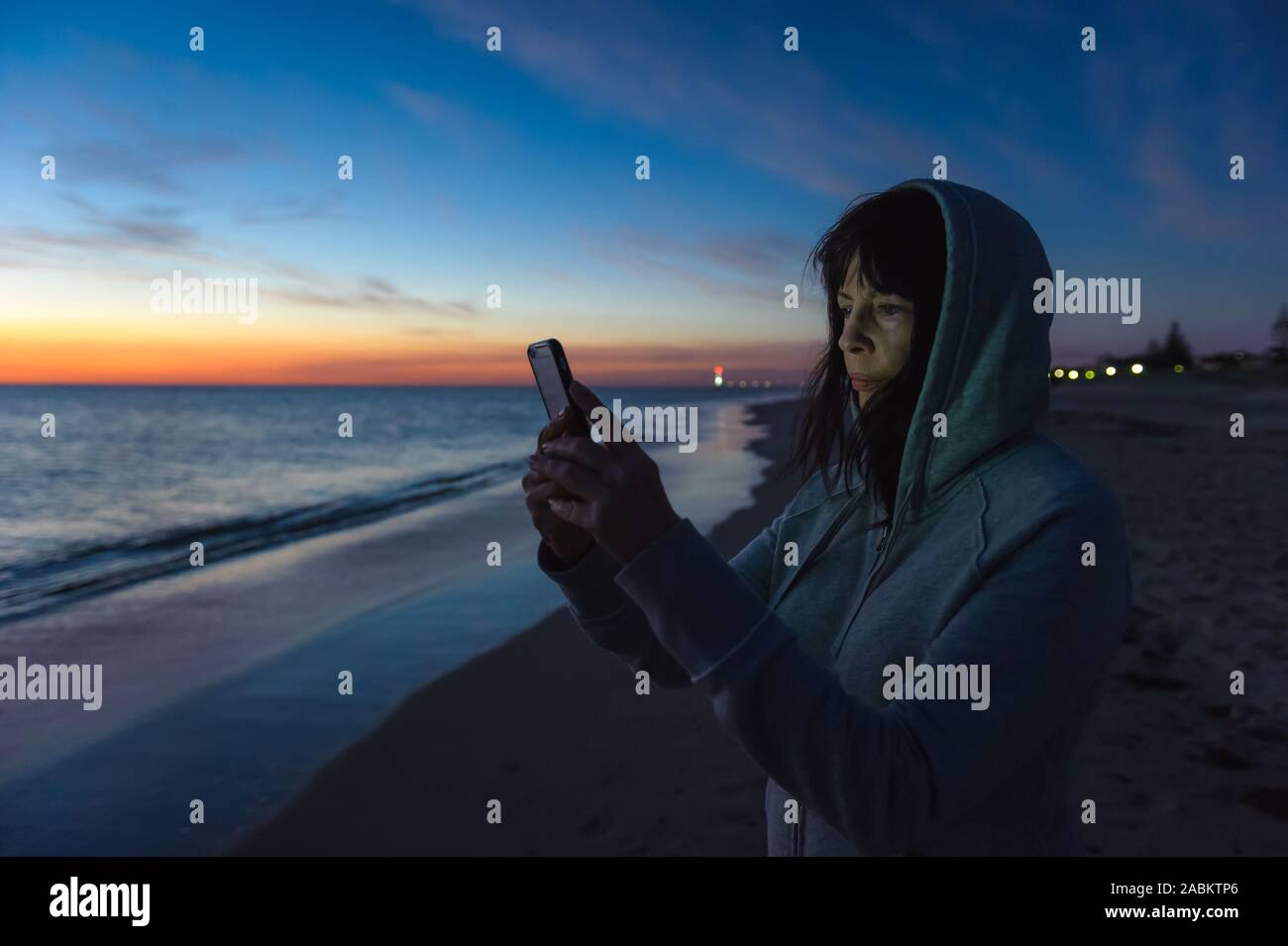 Touristische nach Südaustralien, unterstützt durch eine glorreiche Brighton Beach Sonnenuntergang ihr Gesicht von ihrem Mobiltelefon beleuchtet. Stockfoto