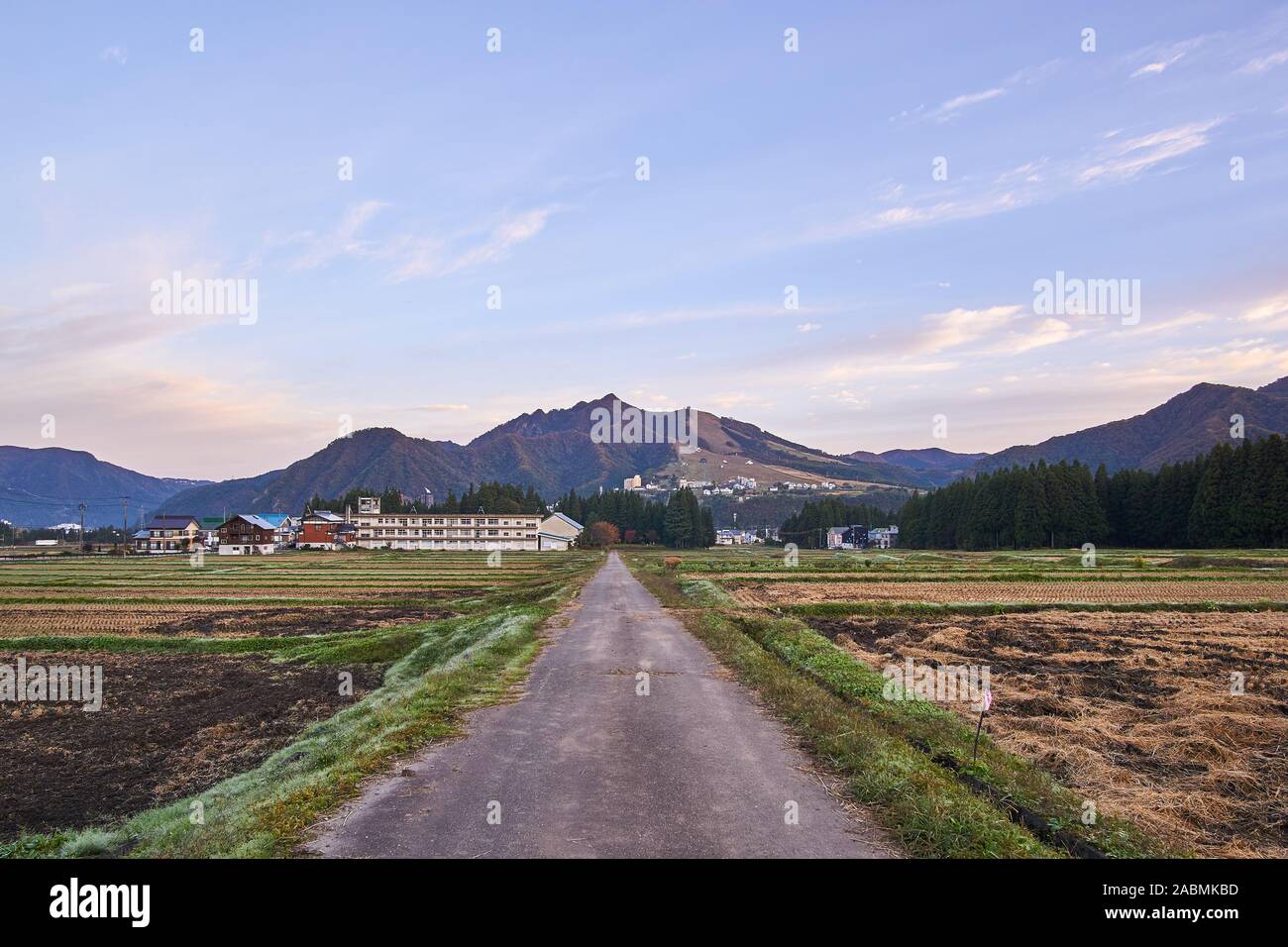 Ein Landstraßen in der Dämmerung an einem klaren Abend führt zu einer Grundschule, Berge, und abgeerntete Reisfelder auf Tsuchitaru, Yuzawa, Niigata, Japan. Stockfoto