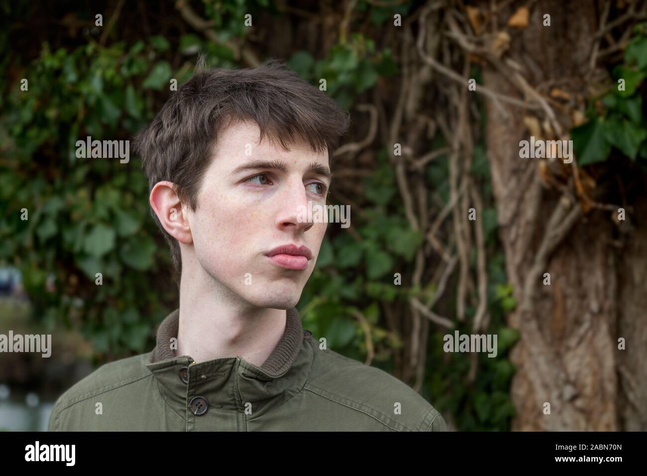 Ein junger Mann, der späten Teens oder 20 etwas steht vor etwas Grün zu seiner Seite nachdenklich. Stockfoto