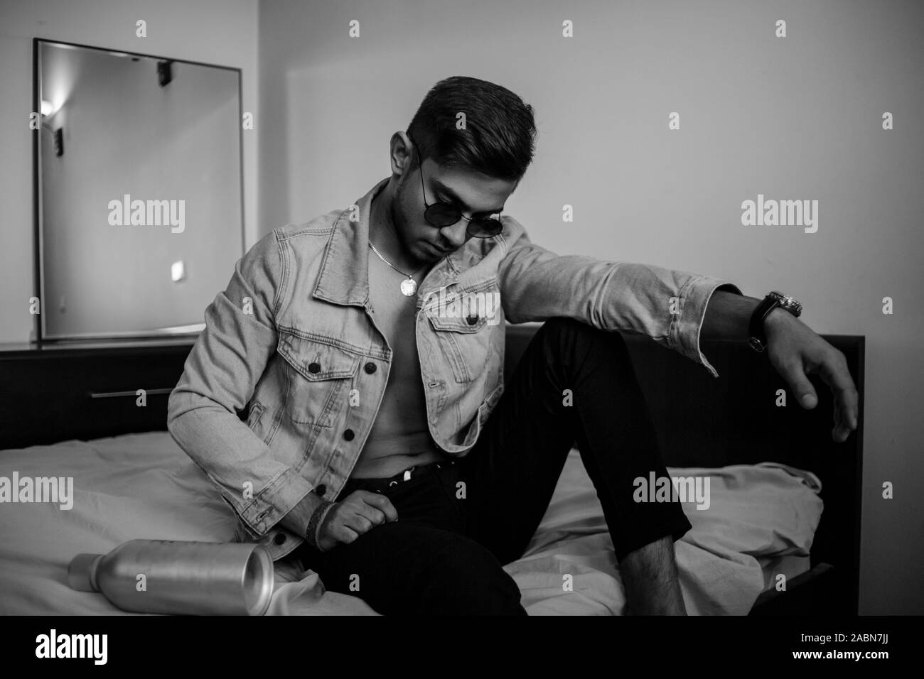 Eine junge stilvolle Kerl in einer Jeansjacke auf dem Bett mit einer Flasche Alkohol. schwarz-weiß Foto mit feinen Korn Wirkung. selektive Fokus Stockfoto