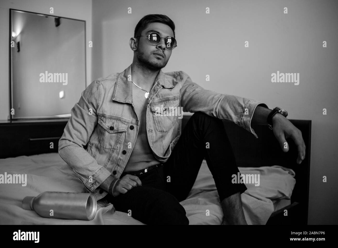 Eine junge stilvolle Kerl in einer Jeansjacke auf dem Bett mit einer Flasche Alkohol. schwarz-weiß Foto mit feinen Korn Wirkung. selektive Fokus Stockfoto