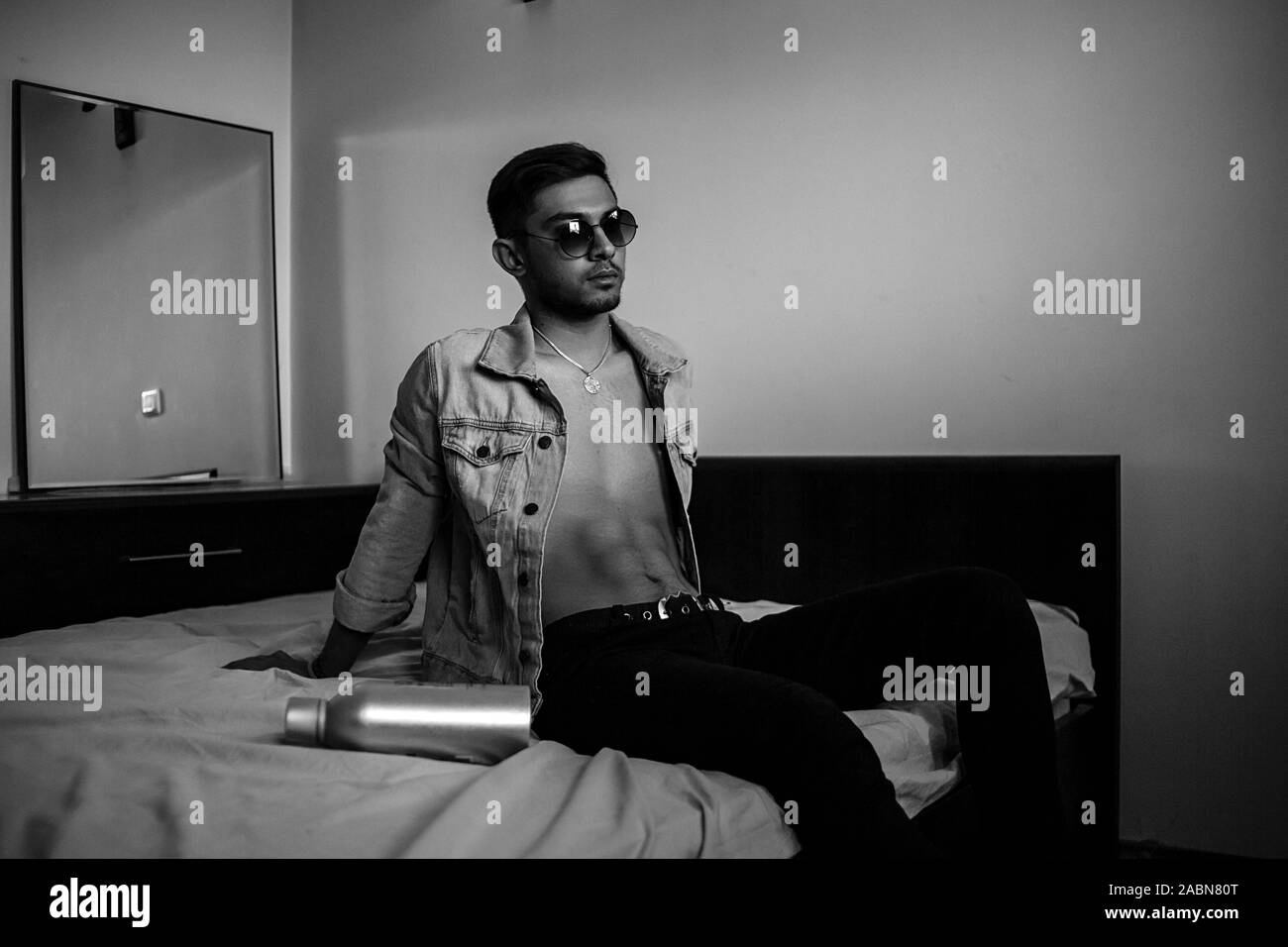 Eine junge stilvolle Kerl in einer Jeansjacke auf dem Bett mit einer Flasche Alkohol. schwarz-weiß Foto mit feinen Korn Wirkung. selektive Fokus Stockfoto