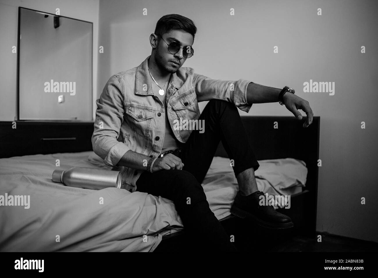 Eine junge stilvolle Kerl in einer Jeansjacke auf dem Bett mit einer Flasche Alkohol. schwarz-weiß Foto mit feinen Korn Wirkung. selektive Fokus Stockfoto