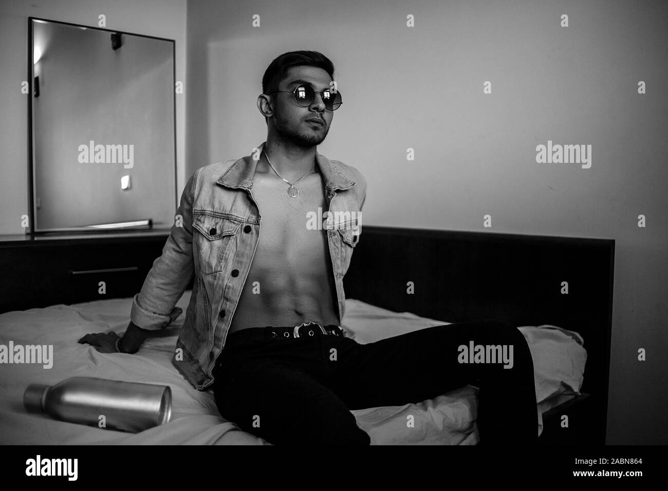 Eine junge stilvolle Kerl in einer Jeansjacke auf dem Bett mit einer Flasche Alkohol. schwarz-weiß Foto mit feinen Korn Wirkung. selektive Fokus Stockfoto