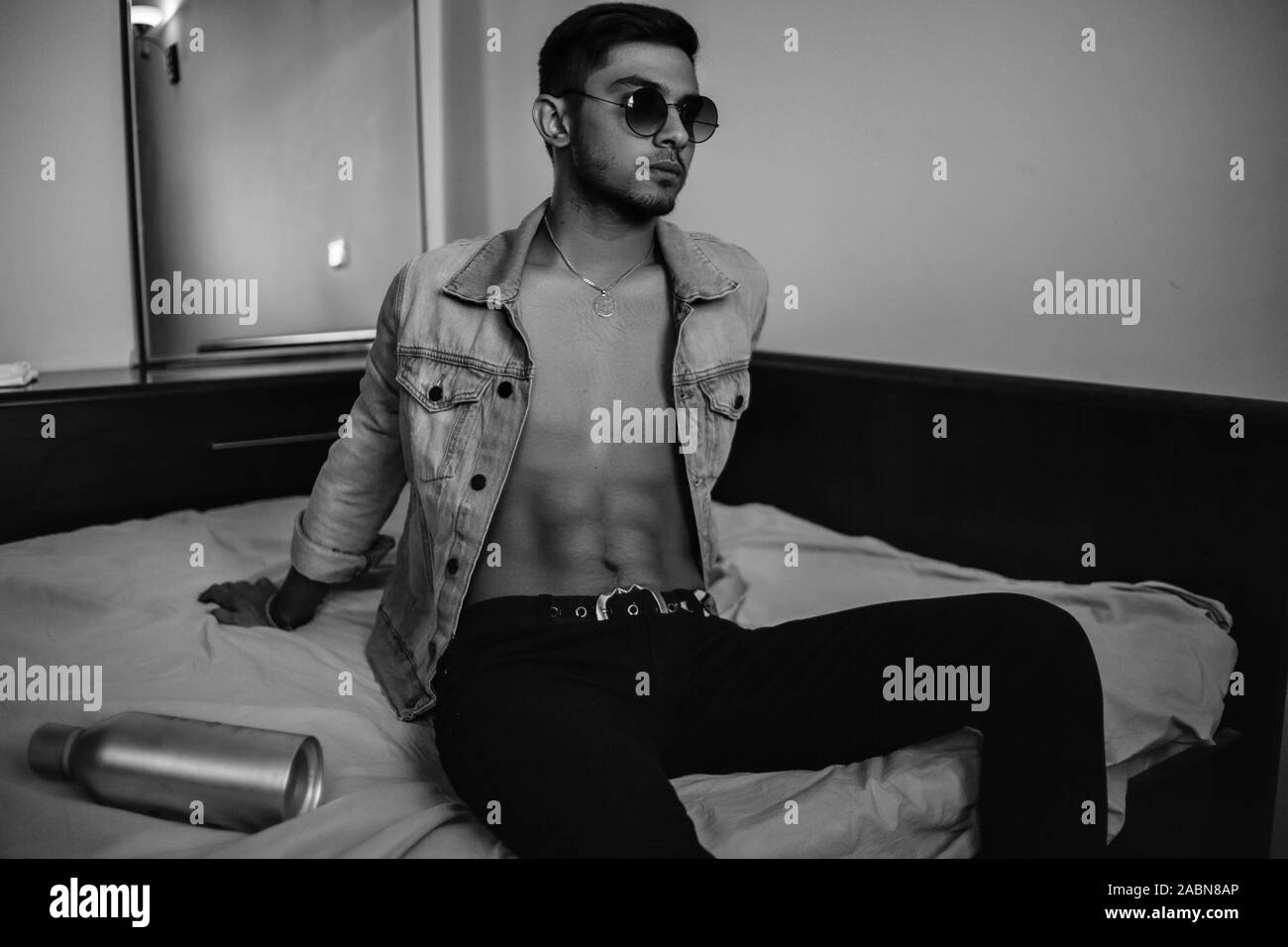 Eine junge stilvolle Kerl in einer Jeansjacke auf dem Bett mit einer Flasche Alkohol. schwarz-weiß Foto mit feinen Korn Wirkung. selektive Fokus Stockfoto