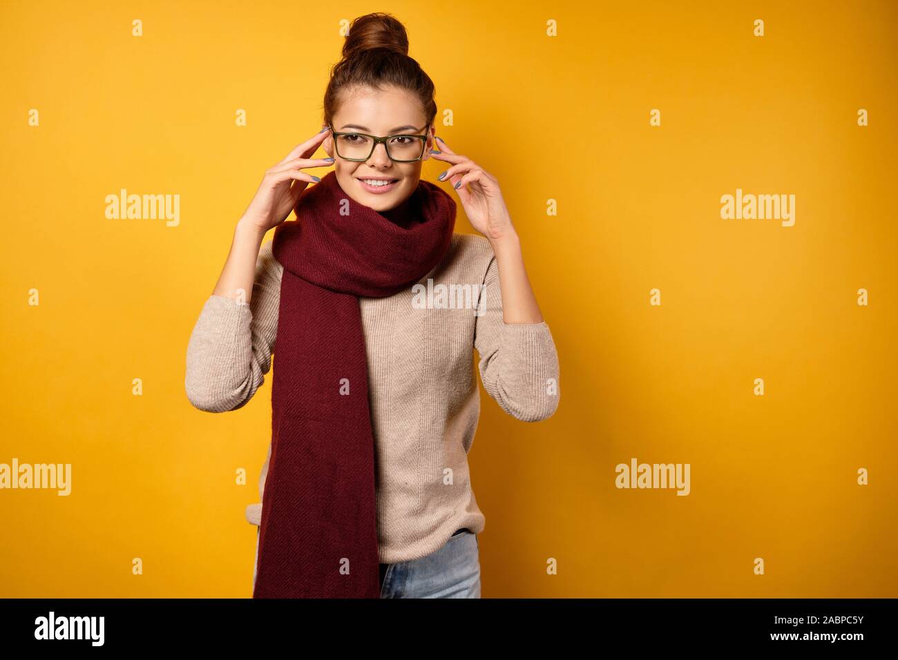 Eine Brünette in einem Schal mit Fernlicht steht auf einem gelben Hintergrund ihre Brille anpassen. Stockfoto