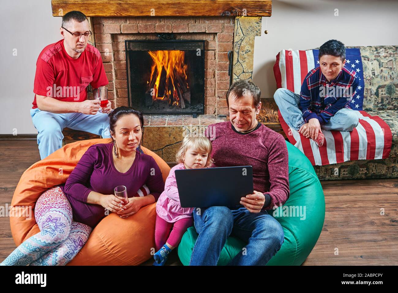 Große Familie verbringen die Zeit zusammen in der Nähe von Kamin: Erwachsene sind, Wein trinken und auf den Bildschirm des Notebooks beim Sitzen auf dem Sitzsack, Stockfoto