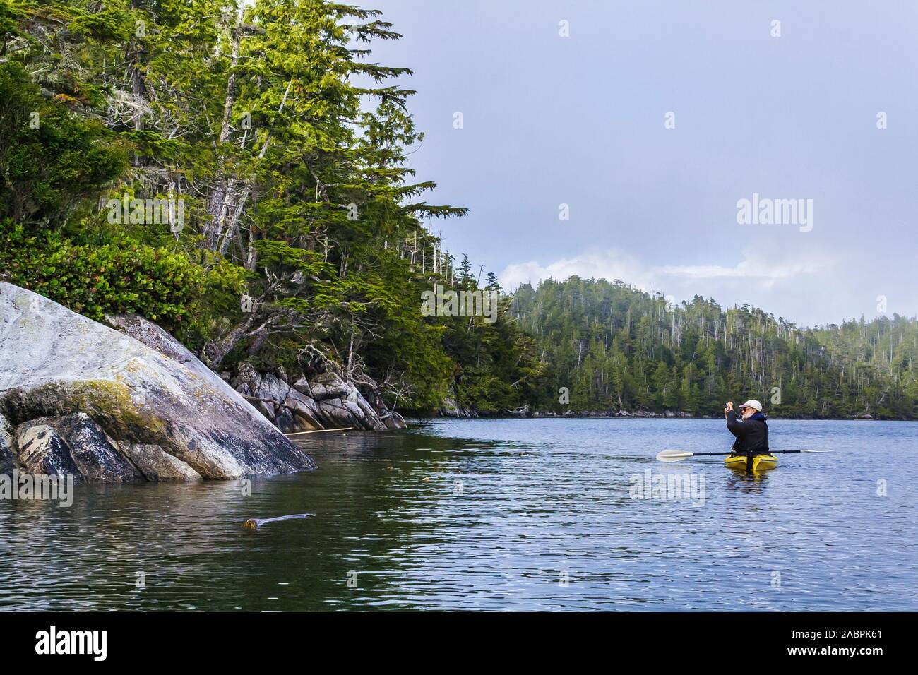 Ein einsamen Mann mittleren Alters schwebt in einem Kajak, hält eine Kamera hoch, um eine felsige, bewaldete Küste in einem abgelegenen Gebiet der Küste von British Columbia zu fotografieren. Stockfoto