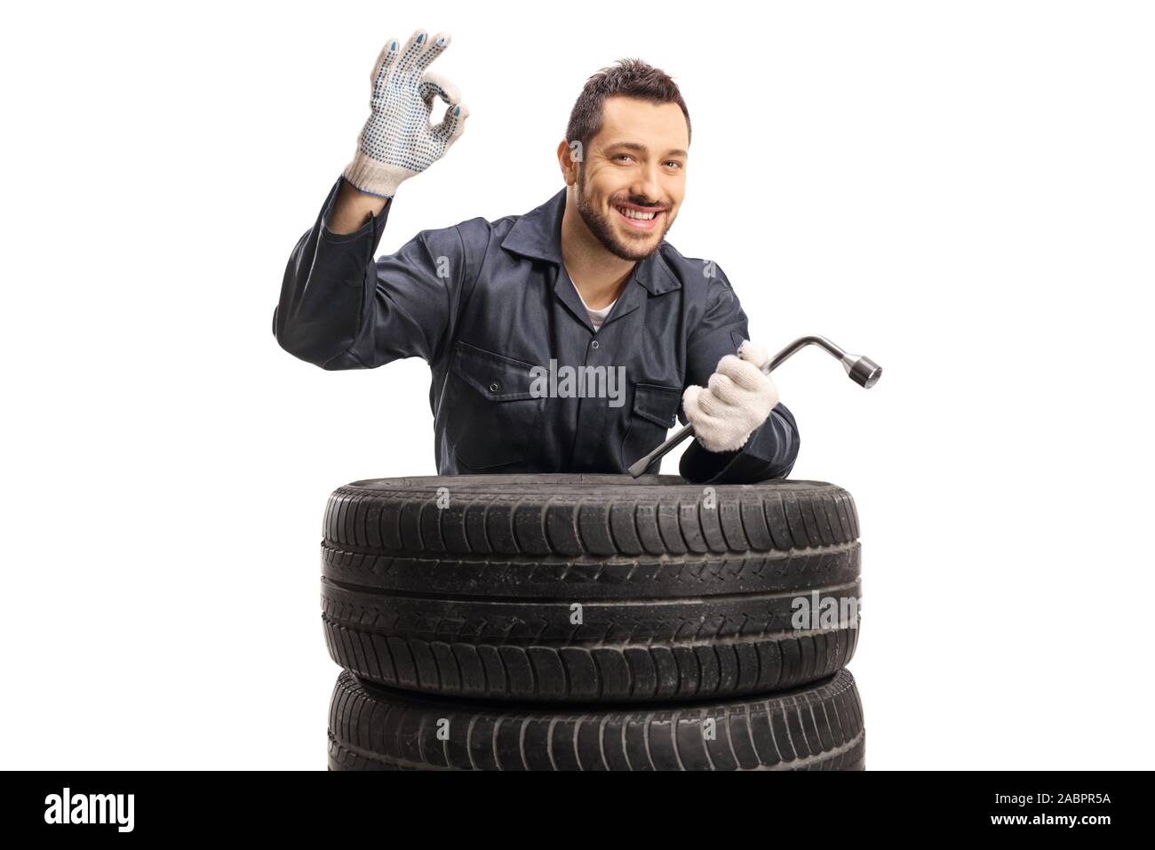 Fröhliche Automechaniker Gestik großartige mit Hand und hält Schraubenschlüssel hinter dem Auto Reifen auf weißem Hintergrund Stockfoto