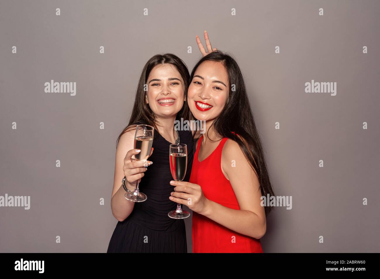 Junge Frauen tragen Abendkleider Studio stehen auf grauen Hintergrund halten Gläser Champagner posiert für die Kamera angezeigt Gruß Geste l isoliert Stockfoto