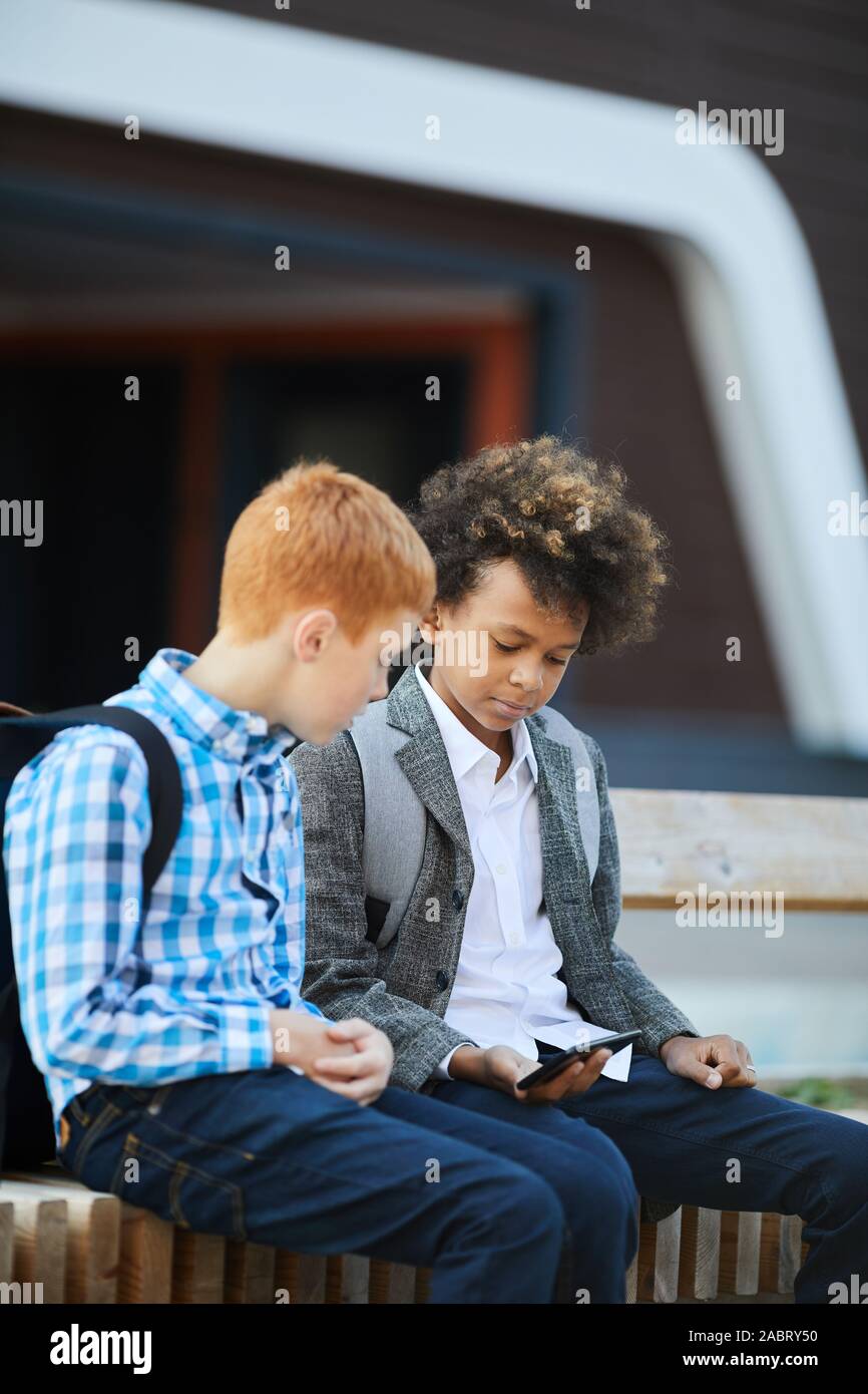 Zwei Jungen in Freizeitkleidung sitzen und über Handy zusammen nach der Schule im Freien Stockfoto