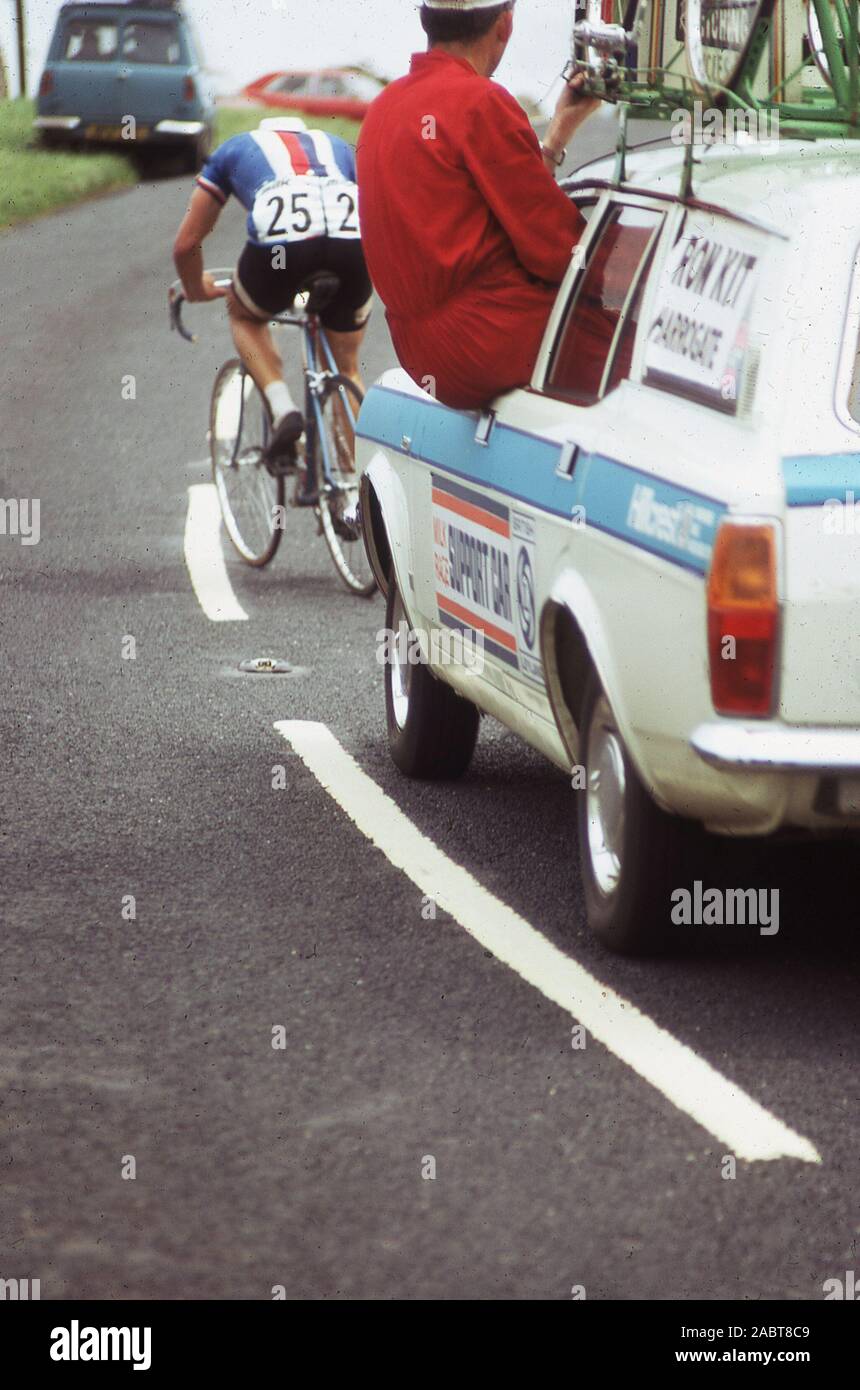 1960er Jahre, historisch, bei einem Radrennrennen, hinter einem Rennradfahrer, einem Mechaniker in roten Overalls, der auf dem Fenstervorsprung eines Hilfswagens sitzt, bereit ist, dem Radfahrer zu helfen, der auf der Straße vor uns kämpft, England, Großbritannien. Stockfoto