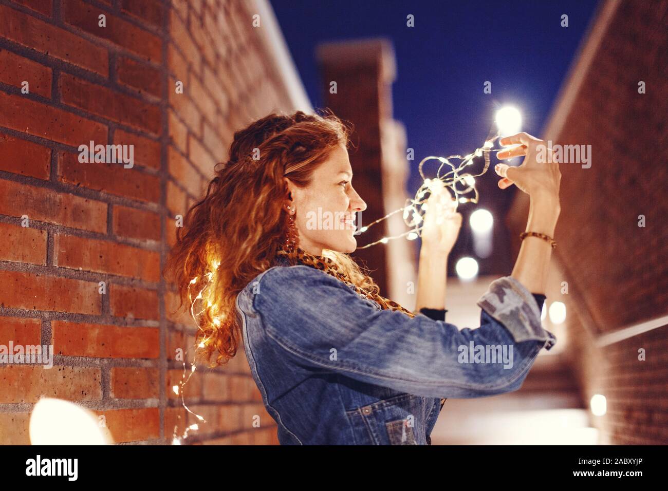 Junge fröhliche Rothaarige kaukasische Frau spielen mit Lichterketten im Freien Stockfoto