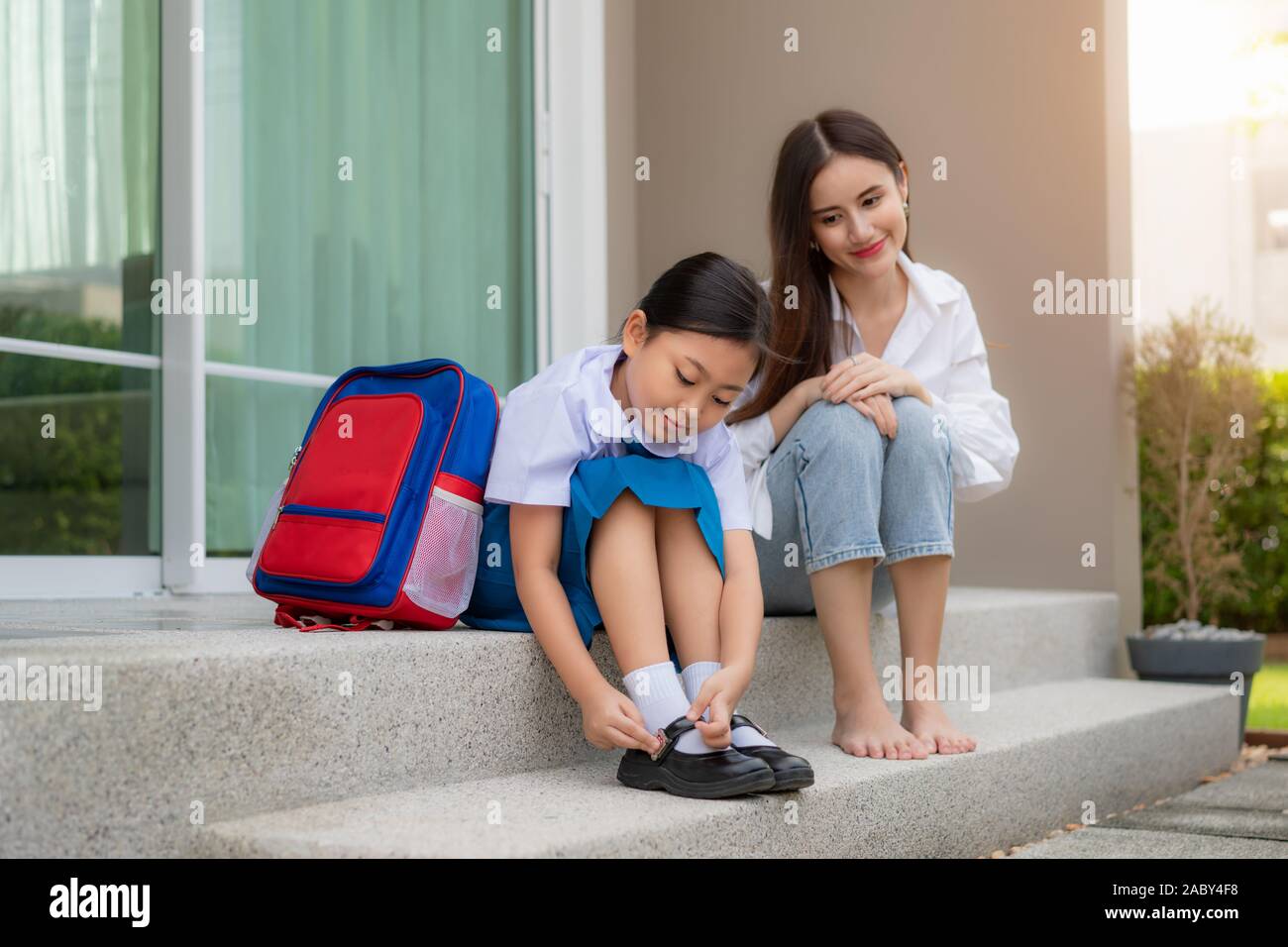 Asiatische Mutter gerade ihre Tochter Vorschule in Uniform zu Ihren eigenen Schuhen infront von zu Hause für die Schule in das schöne Wetter vorbereiten Stockfoto