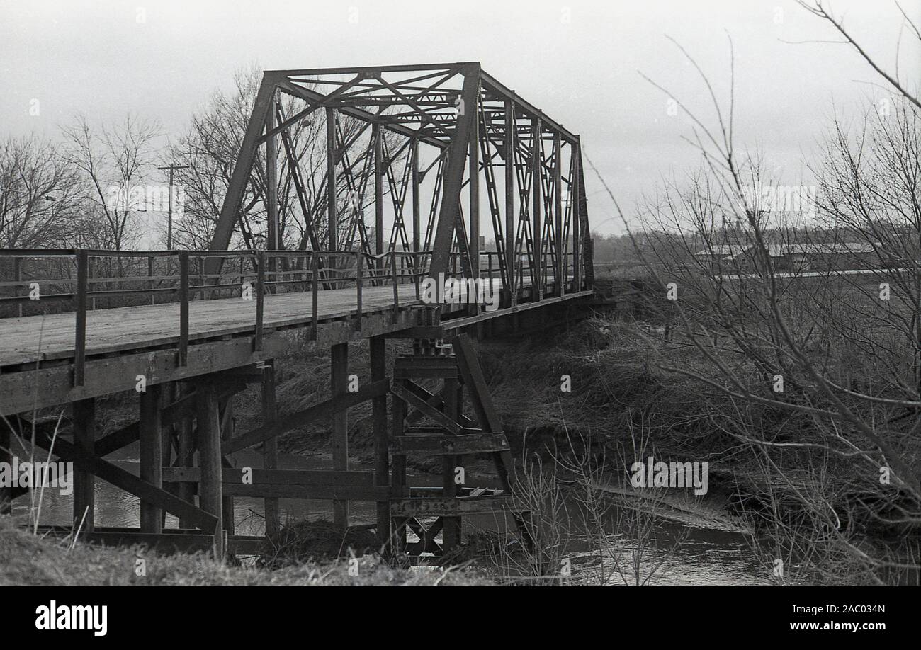 1980er Jahre, historisch, eine hölzerne Brücke mit Metall umschlossenen Abschnitt oder über einen kleinen Fluss im ländlichen mittleren Westen Amerikas. Die ersten als Fachwerkbrücken bekannten Fachwerkbrücken verwendeten Holzbalken als Hauptdeckkonstruktion mit zusätzlichen Stahlstreben, die in dreieckiger Form gruppiert waren, um die Brücke zu stützen. Fachwerkbrücken gibt es in vielen verschiedenen Ausführungen. Stockfoto