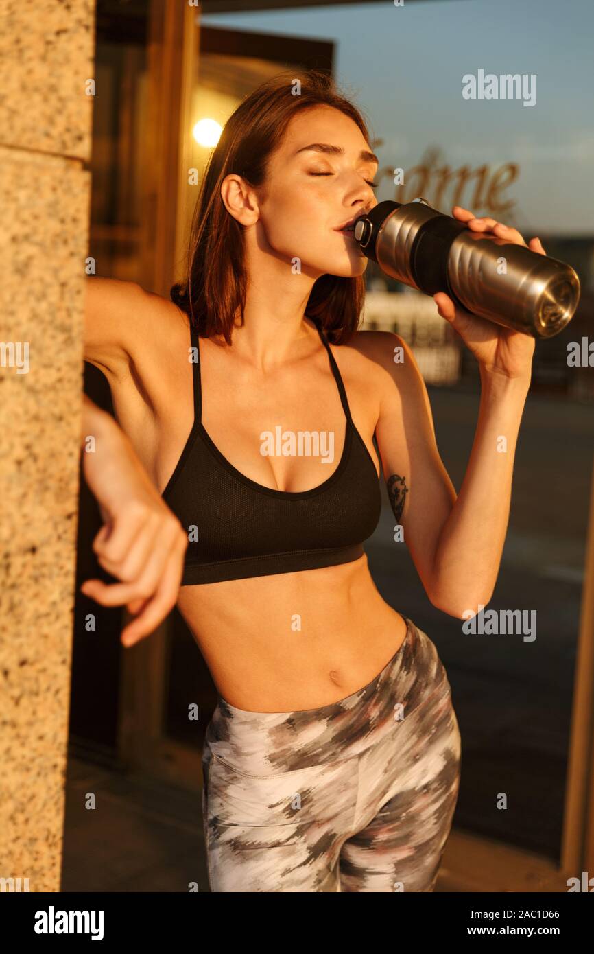 Image der Jungen schlanken Frau in Sportswear Trinkwasser aus der Flasche, während Sie in der Nähe von Gebäude arbeiten in Morgen Stockfoto