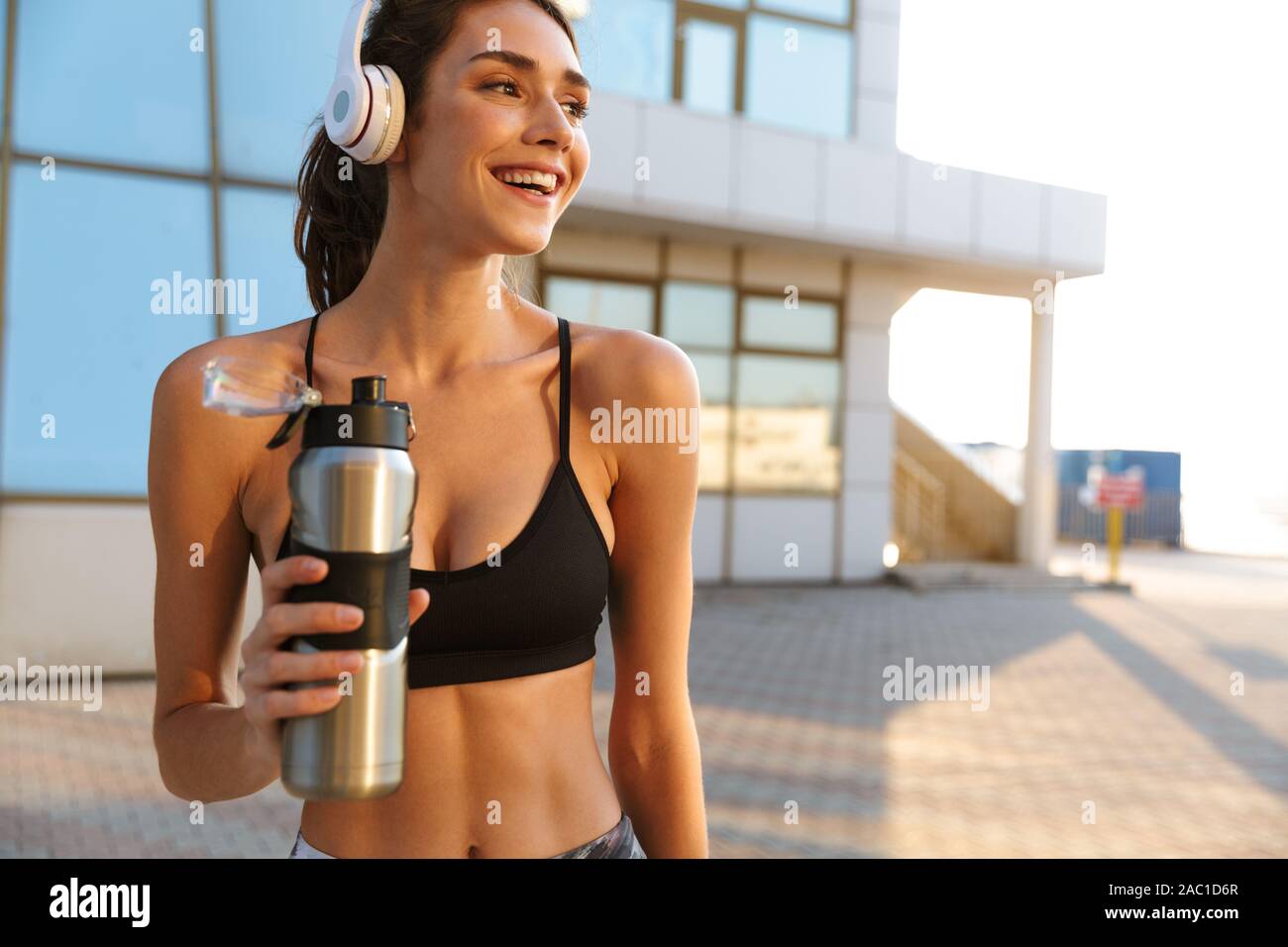 Bild lächeln gerne Frau in Sportkleidung mit Kopfhörern und Trinkwasser, während Sie in der Nähe von Glas Gebäude arbeiten in Morgen Stockfoto