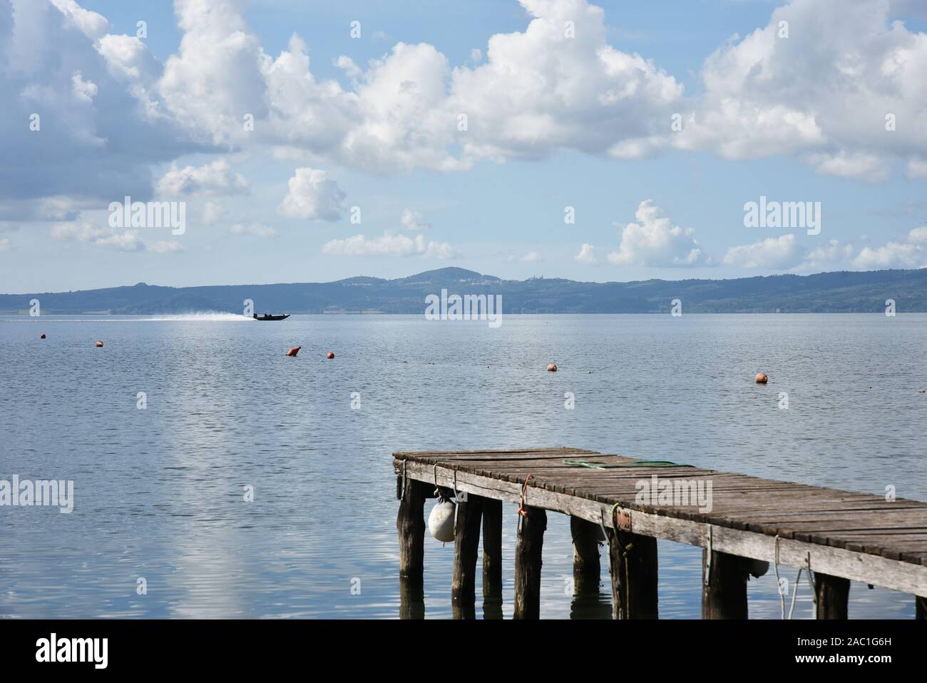 Hölzerne Anlegesteg für Boote vor blauem Himmel mit einigen Wolken. Bojen. Hügeln im Hintergrund. Bolsena See, Italien. Stockfoto