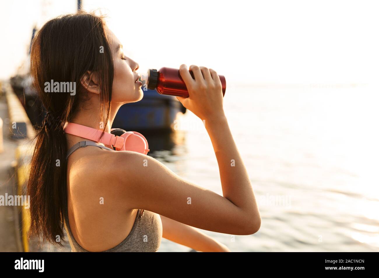 Bild des athletischen asiatische Frau in Sportliche Kleidung mit Kopfhörern Trinkwasser bei einem Spaziergang am Meer port Stockfoto
