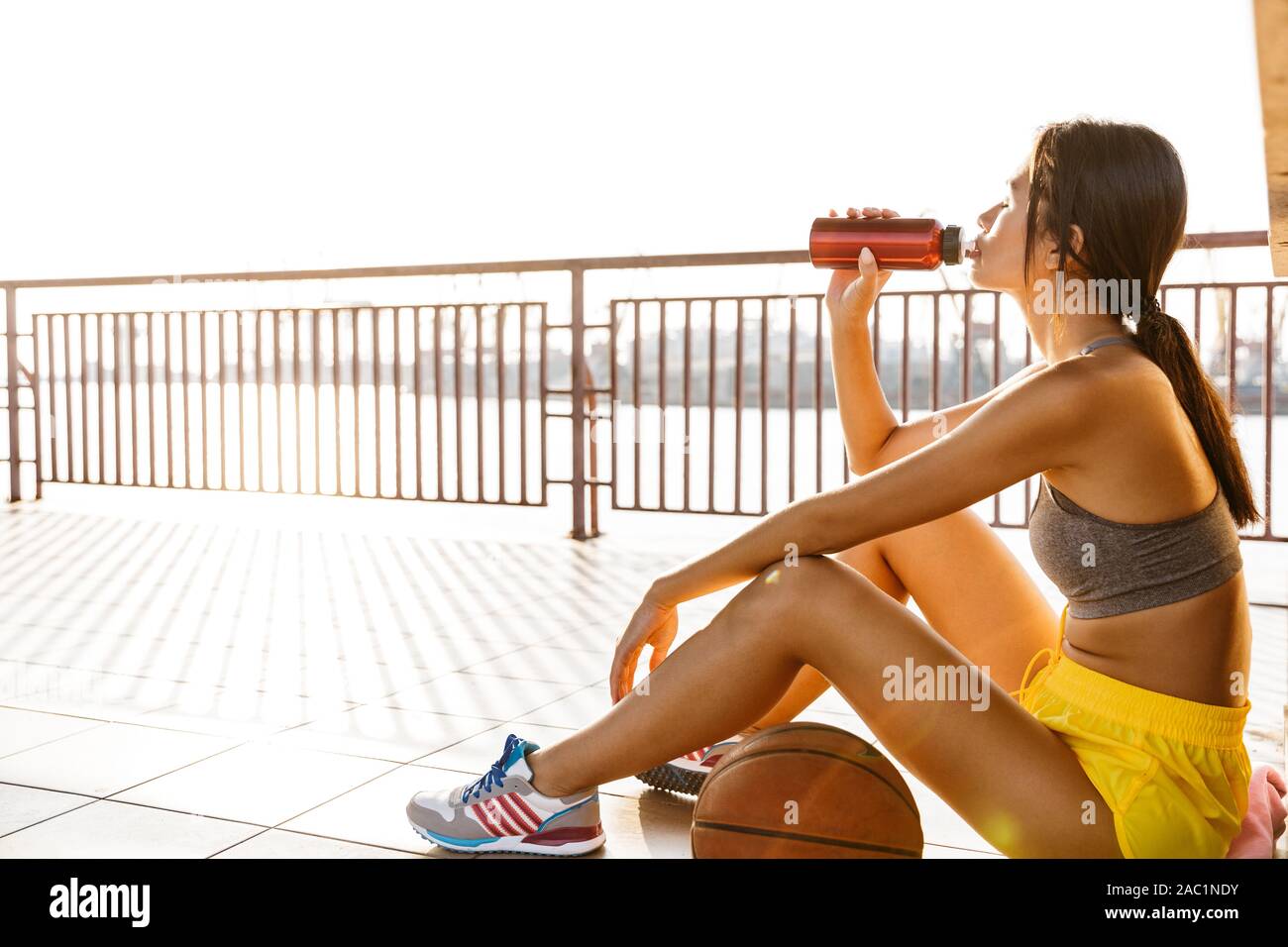Bild von Fitness asiatische Frau in sportlicher Bekleidung Trinkwasser beim Sitzen mit Basketball am Meer port Stockfoto