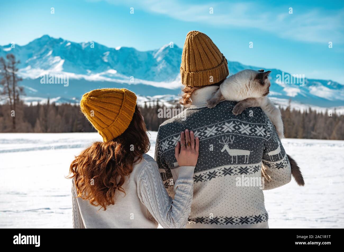 Junges Paar mit Katze auf der Schulter steht in Pullover und Strick Hüte gegen Berge Stockfoto