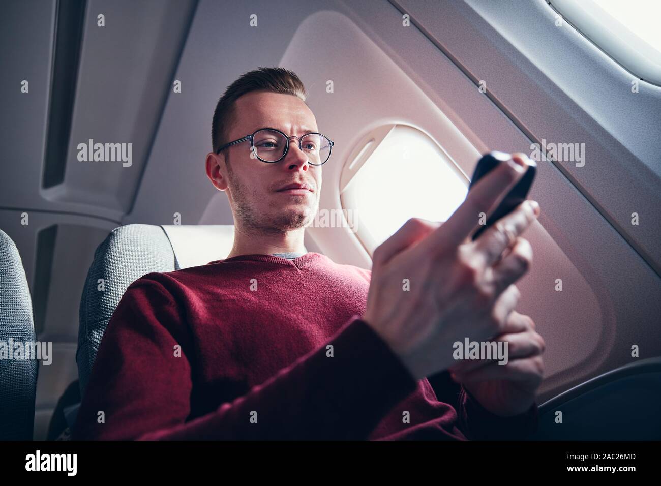 Internetverbindung während des Fluges. Junger Mann mit Brille mit Handy im Flugzeug. Stockfoto