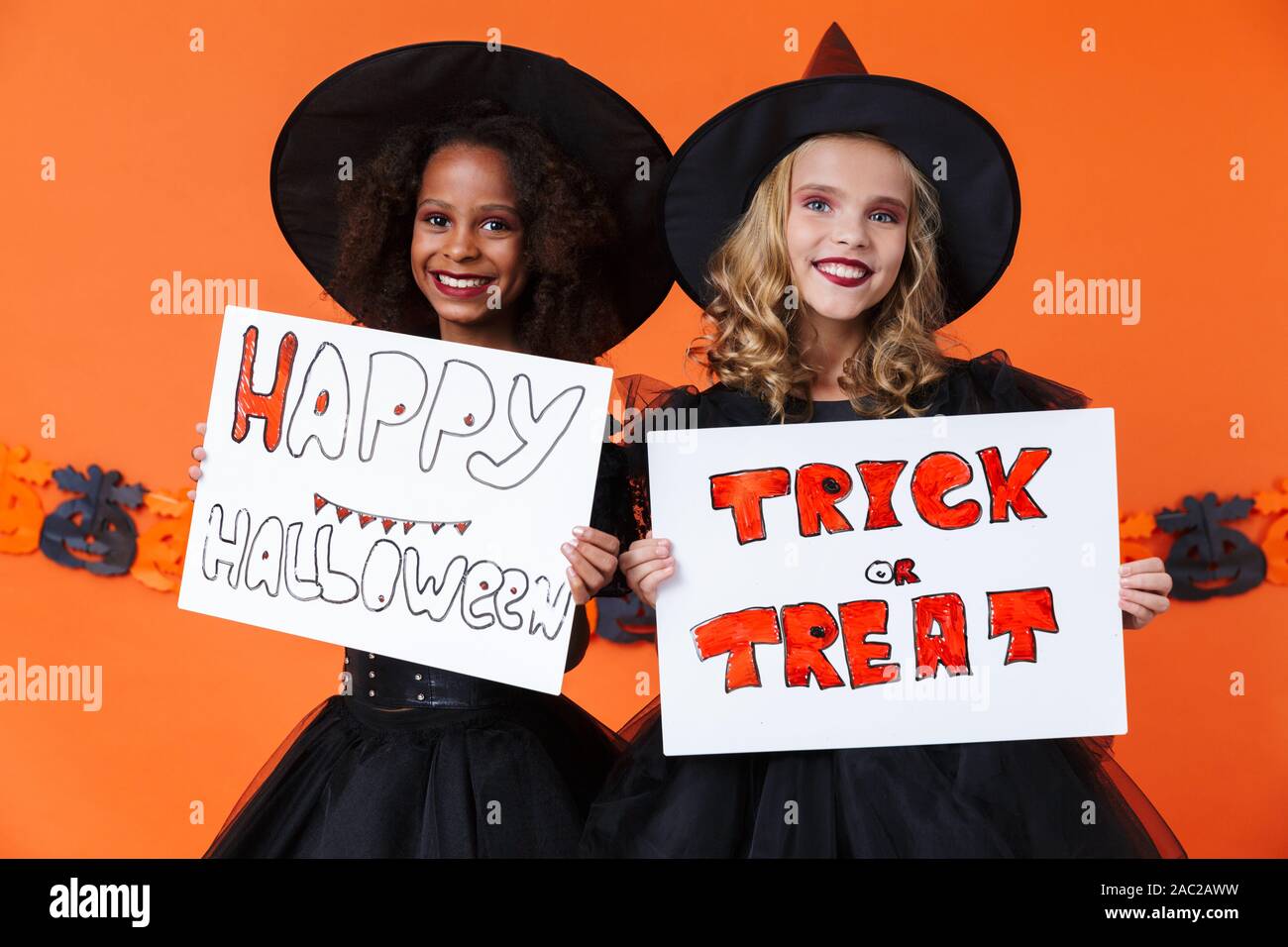 Bild von lächelnden multinationale Mädchen in Schwarz halloween Kostüme Plakate über orange Kürbis Wand isoliert Stockfoto