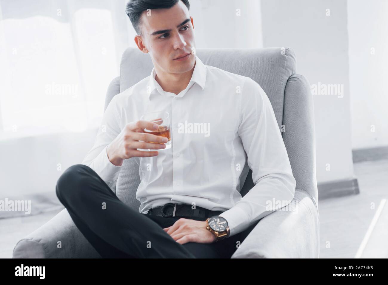 Auf der Seite. Nähe zu sehen. Junge elegante Mann im weissen Anzug auf dem Stuhl und hält Glas mit Alkohol Stockfoto