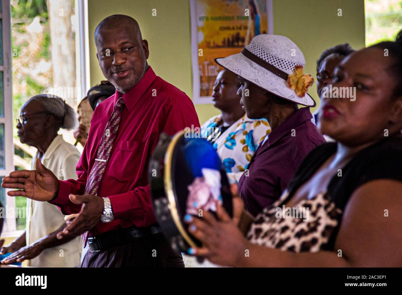 Ein Mann in einem roten Hemd klatscht mit erhobenen Händen während des Baptisten-Gottesdienstes in The Lime, Grenada. Eine Frau mit einem weißen Hut spielt neben ihm eine Tamburin. Der Gesang schafft eine gemeinsame Gemeinschaftserfahrung. Viele Gläubige bringen ihr eigenes Instrument zum Gottesdienst mit. Stockfoto