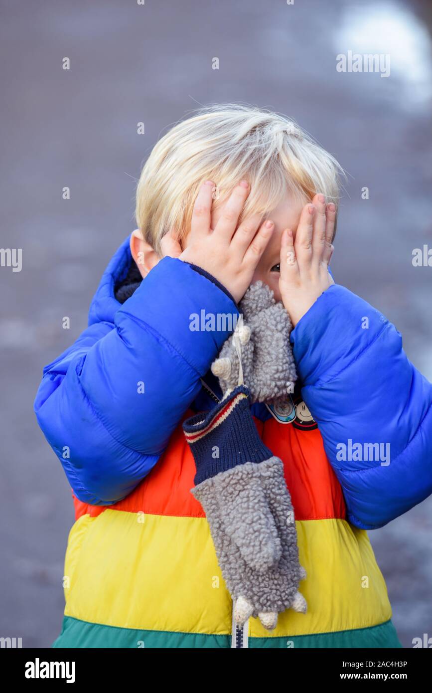 Ein- und Ausblenden der Junge außerhalb spielt in Outdoor Bekleidung ist er für Gesicht und verstecken, während sie in die Kamera schaut suchen Stockfoto