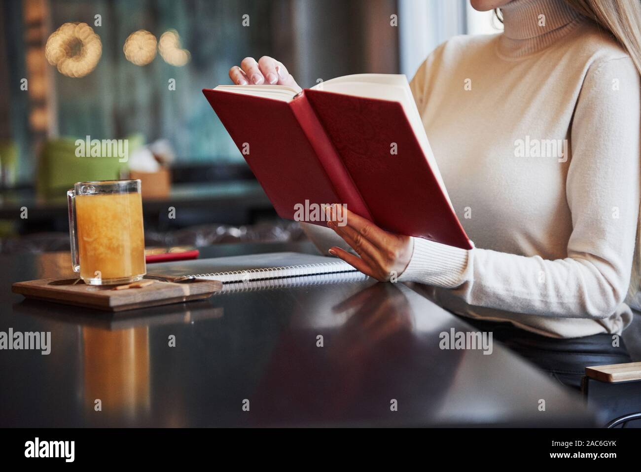 Blonde Mädchen lesen Sie ein Buch mit rotem Deckel im Restaurant mit Tisch und gelbe Getränk auf es Stockfoto