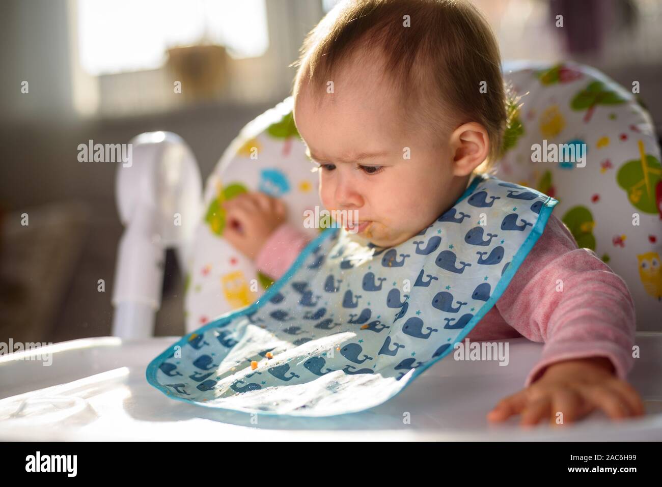 Baby nicht gefallen das Essen und spuckt es aus. Das Sitzen in einem Hochstuhl im hellen Wohnzimmer. 6 - 12 Monate alt. Fütterung baby Konzept Stockfoto