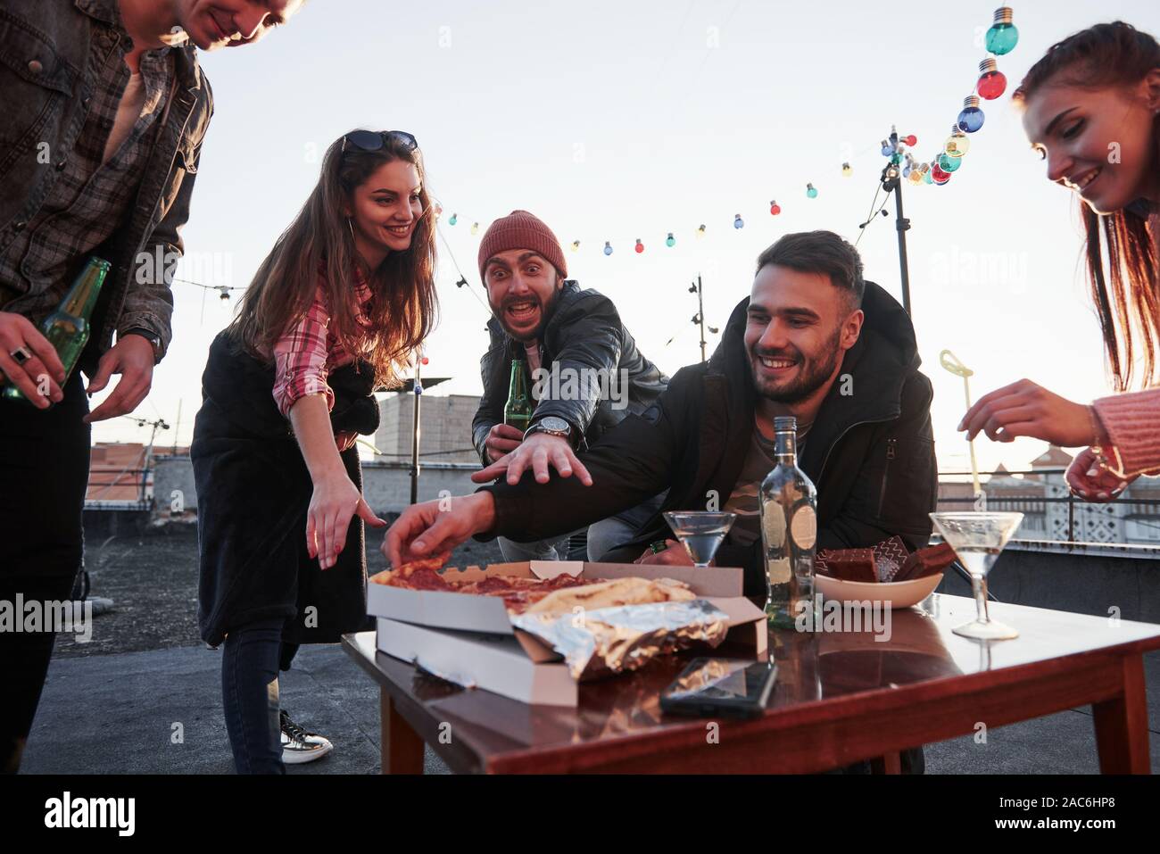 Kerl in der Red Hat macht verrückt hungrige Gesicht und versucht, dass frisches Produkt zu erreichen. Essen Pizza auf der Dachterrasse. Gute Freunde haben Wochenende mit Stockfoto