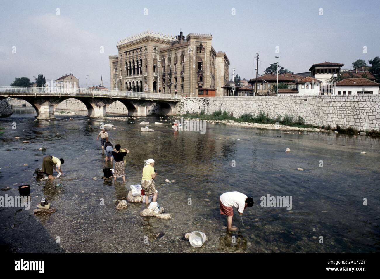 9. August 1993 während der Belagerung von Sarajevo: Frauen waschen Wäsche im Fluss Miljacka, mit dem ausgebrannten National Art Gallery und Bibliothek im Hintergrund. Heute ist die Bibliothek von Sarajevo City Hall. Stockfoto