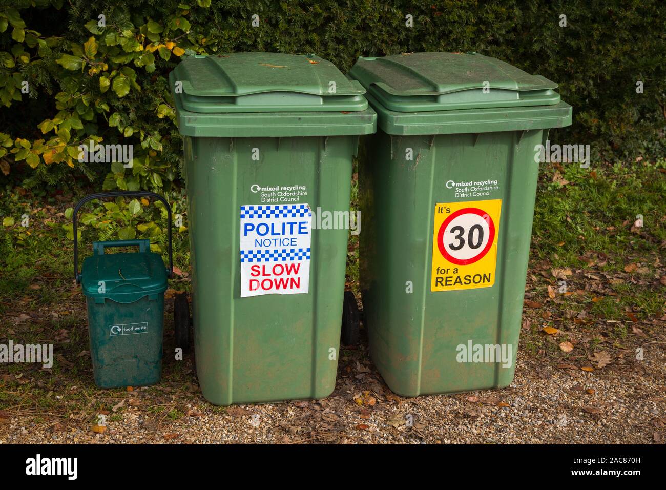 Ein paar grüne Kunststoff Recycling Bins mit höflicher Hinweis keine Beschleunigung der Beschilderung 'Langsamer' und 'Es ist 30 für einen Grund' auf einer Landstraße im Dorf Stockfoto