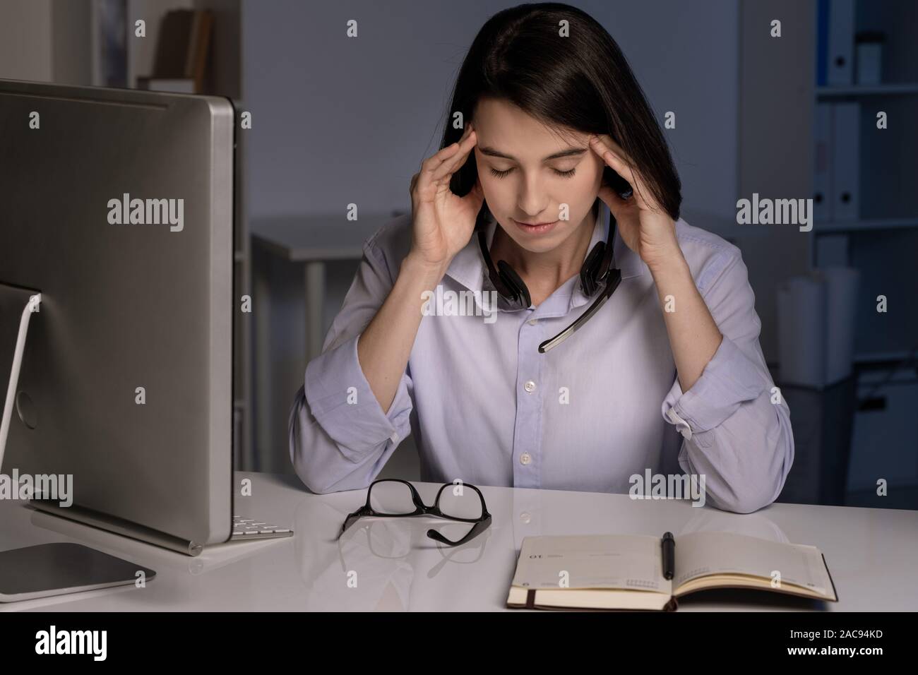 Junge müde Kundendienstmitarbeiter Kopf berühren, während Sie von Arbeitsplatz sitzen am späten Abend Stockfoto