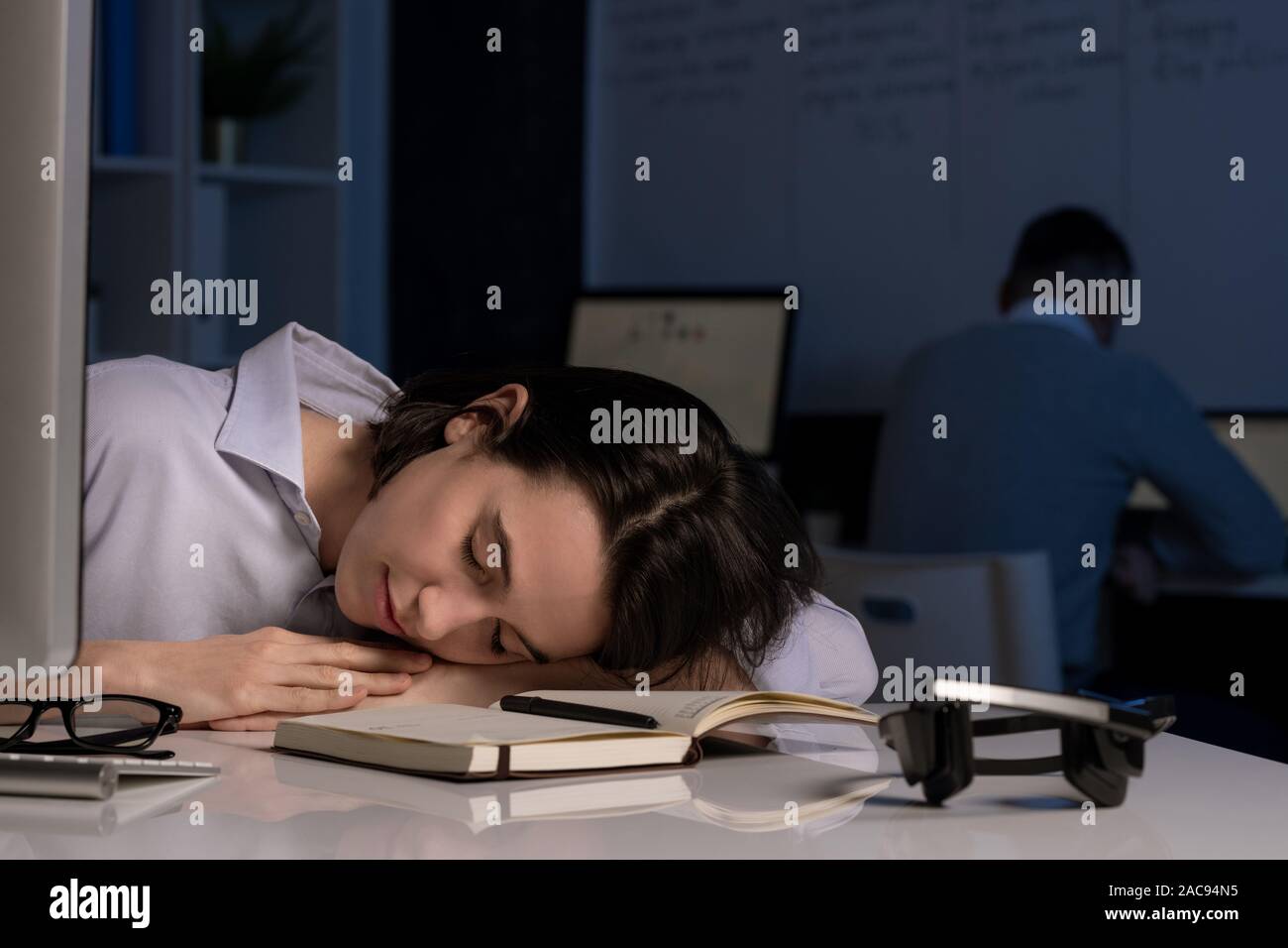 Erschöpft junge weibliche hotline Betreiber napping mit dem Kopf auf dem Schreibtisch nach Arbeiten am späten Abend mit Kollegen im Hintergrund Stockfoto