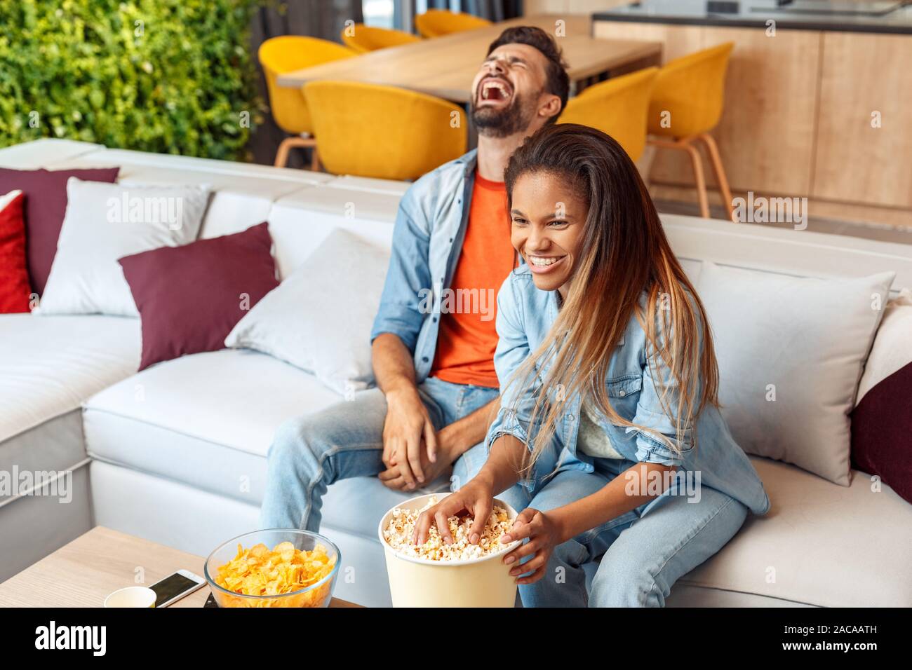 Mischlinge Ehe. Junger Mann und Frau beobachten Komödie im Fernsehen zusammen essen popcorn Lachen heiter Stockfoto