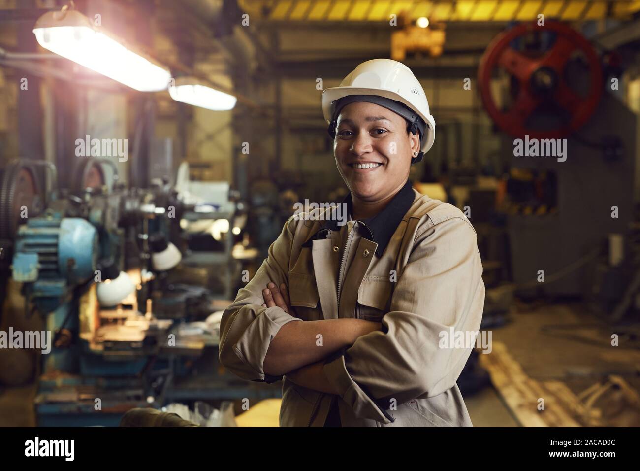 Taille bis Portrait von Mixed-race Arbeiterin selbstbewusst posiert im Stehen mit der Waffe in der Werkhalle gekreuzt Stockfoto