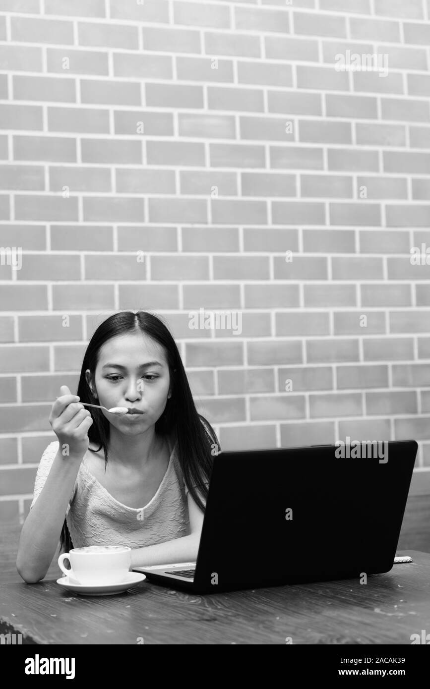 Junge schöne asiatische teenage Mädchen bläst den Schaum des Cappuccino mit Laptop auf hölzernen Tisch gegen die Wand Stockfoto